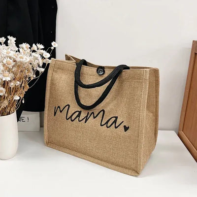 Sac en toile de jute avec inscription "mama", poignées noires, posé sur une table blanche à côté d'un vase de fleurs séchées.