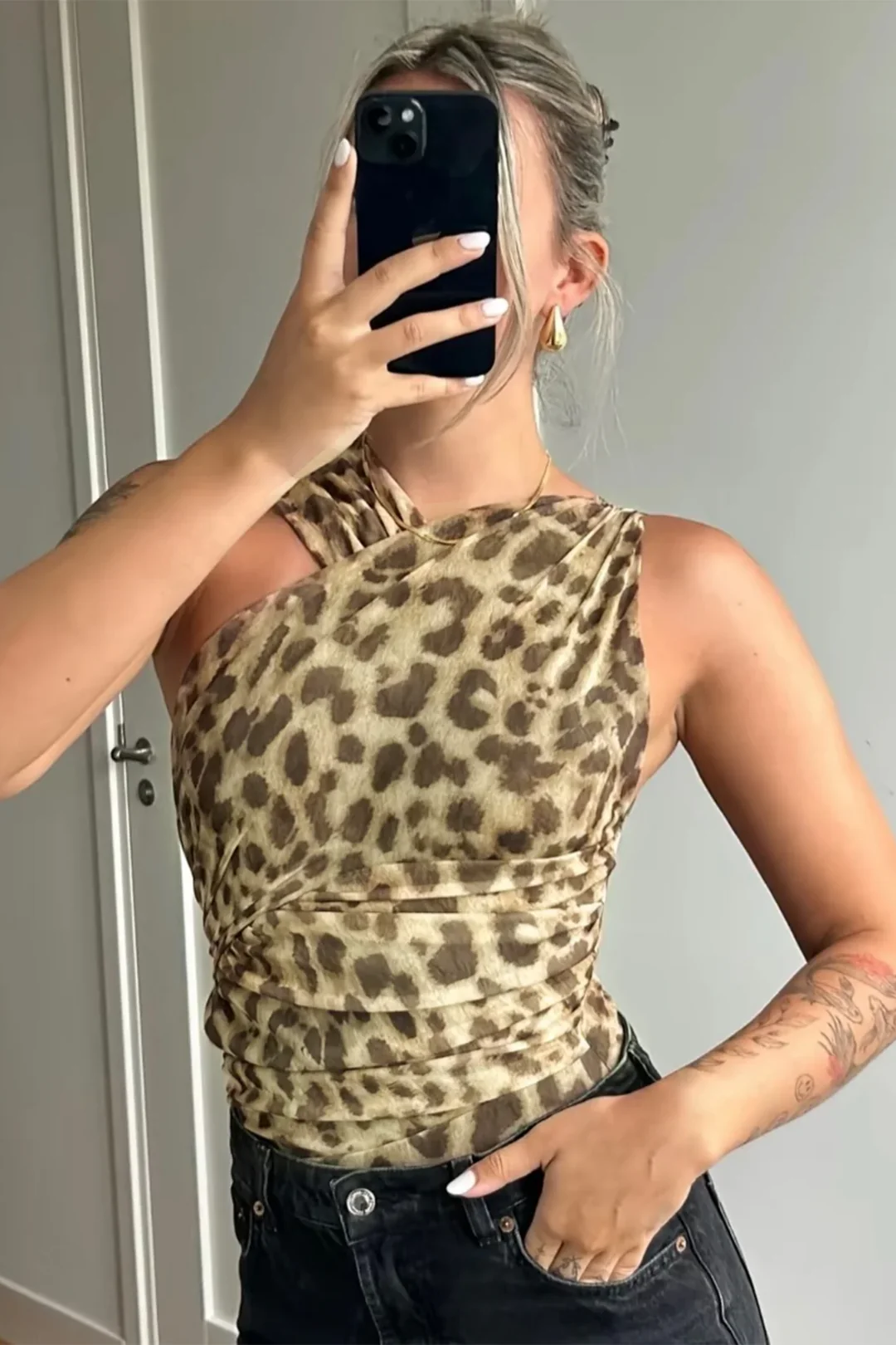 Femme prenant un selfie, portant un haut asymétrique imprimé léopard, cheveux attachés, boucles d'oreilles dorées, tatouages visibles sur le bras.