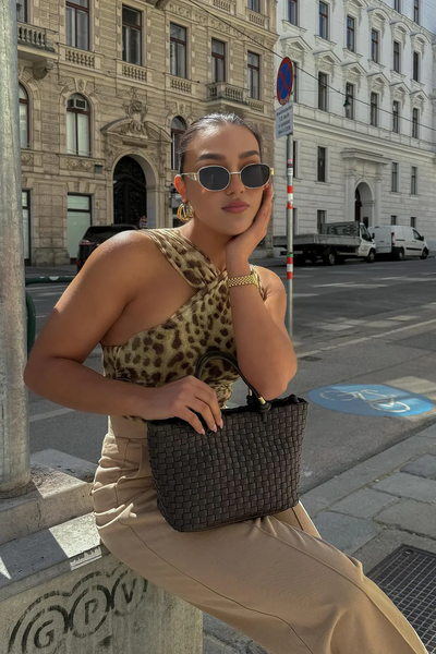 Femme élégante portant un haut léopard et lunettes de soleil, tenant un sac tressé noir, posant dans une rue urbaine avec bâtiments historiques en arrière-plan.