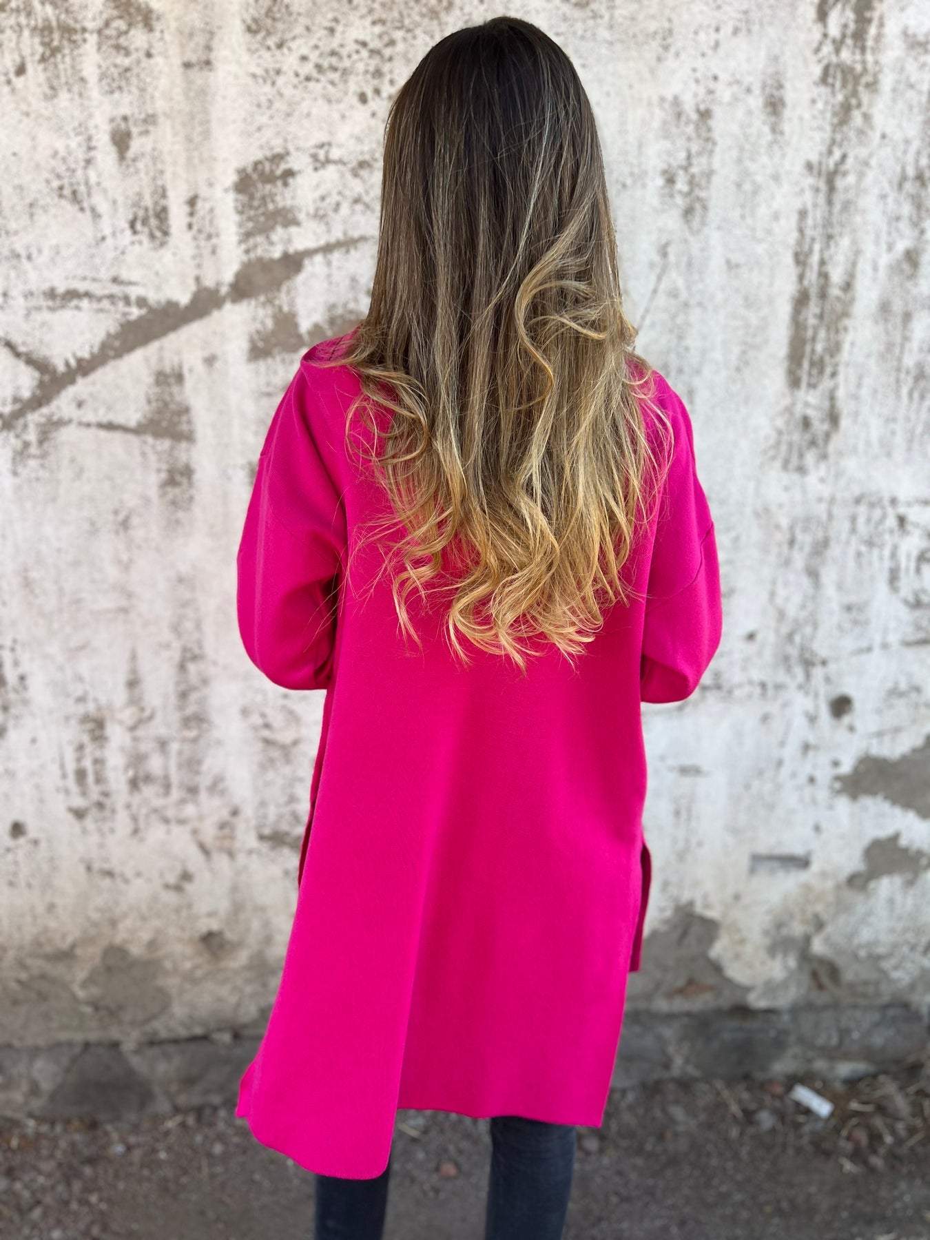 Femme de dos portant un manteau rose vif, cheveux longs et ondulés, devant un mur texturé gris. Mode féminine, style urbain, automne.