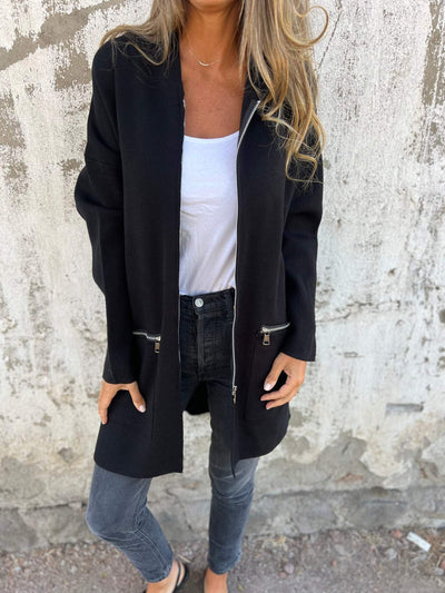 Femme portant un manteau noir élégant avec fermeture éclair, t-shirt blanc, jeans gris, posant devant un mur texturé. Mode automne chic.