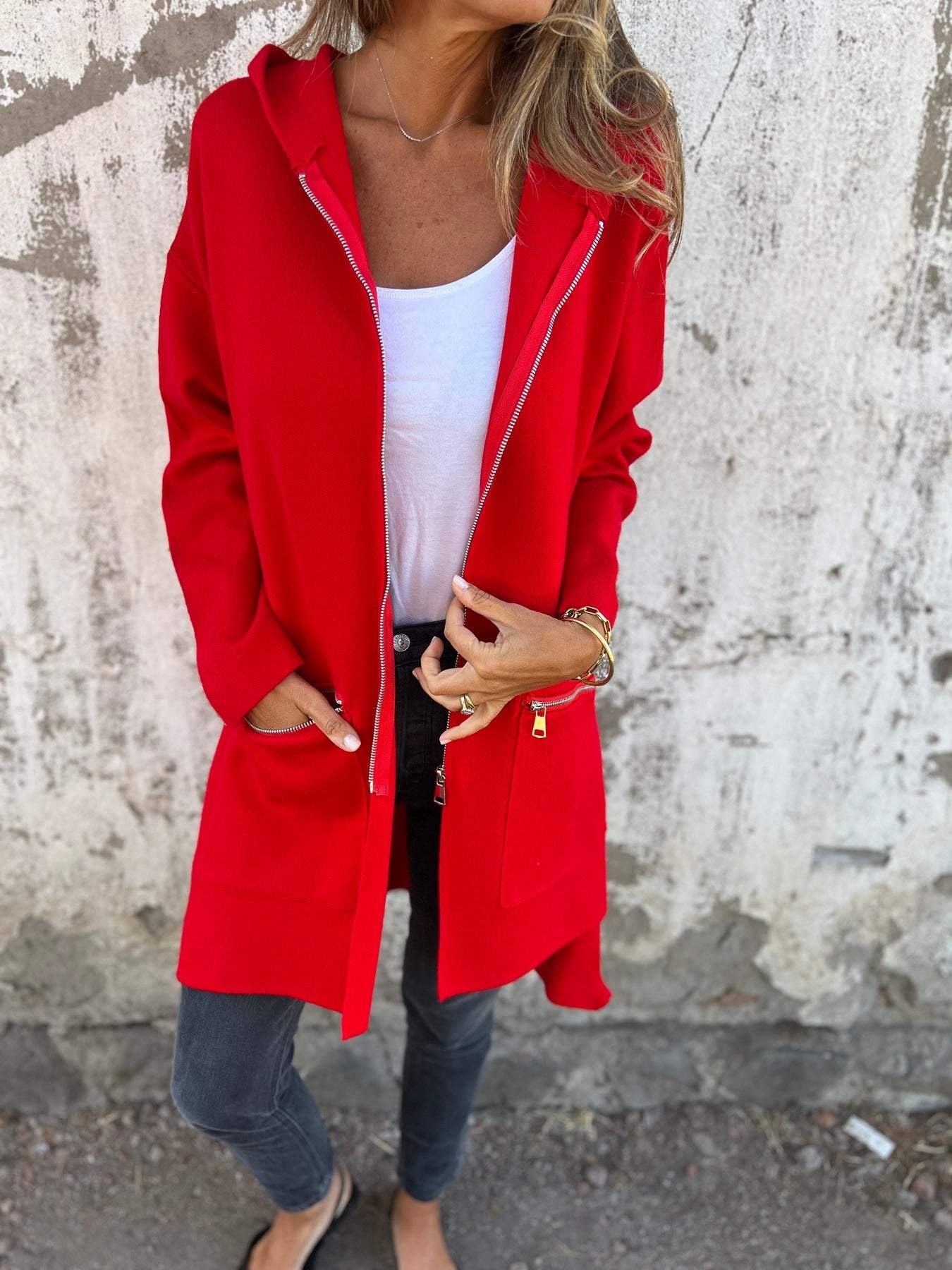 Femme portant un manteau rouge vif avec capuche, t-shirt blanc, jeans noirs, posant devant un mur texturé. Mode automne, style décontracté.