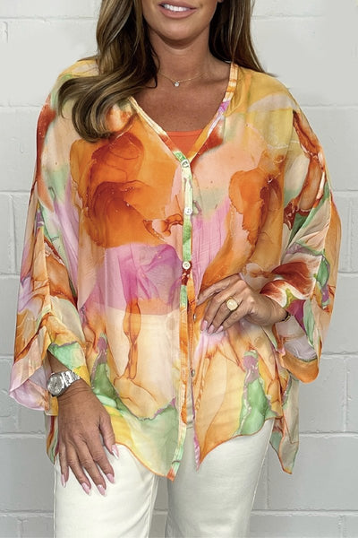 Femme portant une chemise fluide colorée à motifs floraux orange et rose, manches longues, sur fond de mur blanc. Mode féminine, style décontracté.