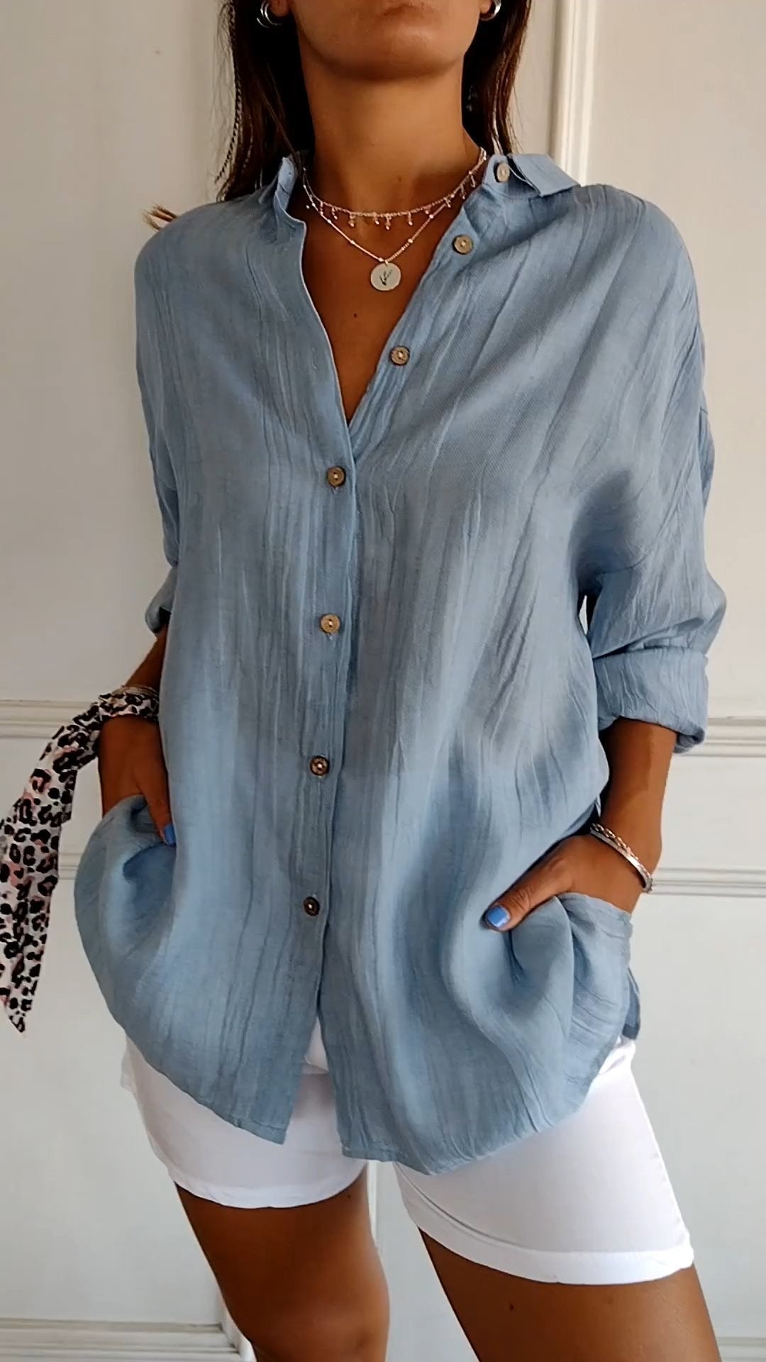 Femme portant une chemise bleue en lin avec boutons en bois, manches retroussées, collier en argent, short blanc; style décontracté et élégant.