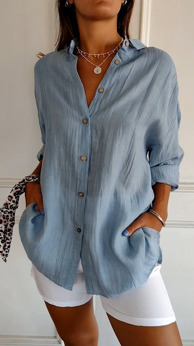 Femme portant une chemise bleue en lin avec boutons en bois, manches retroussées, collier en argent, short blanc; style décontracté et élégant.