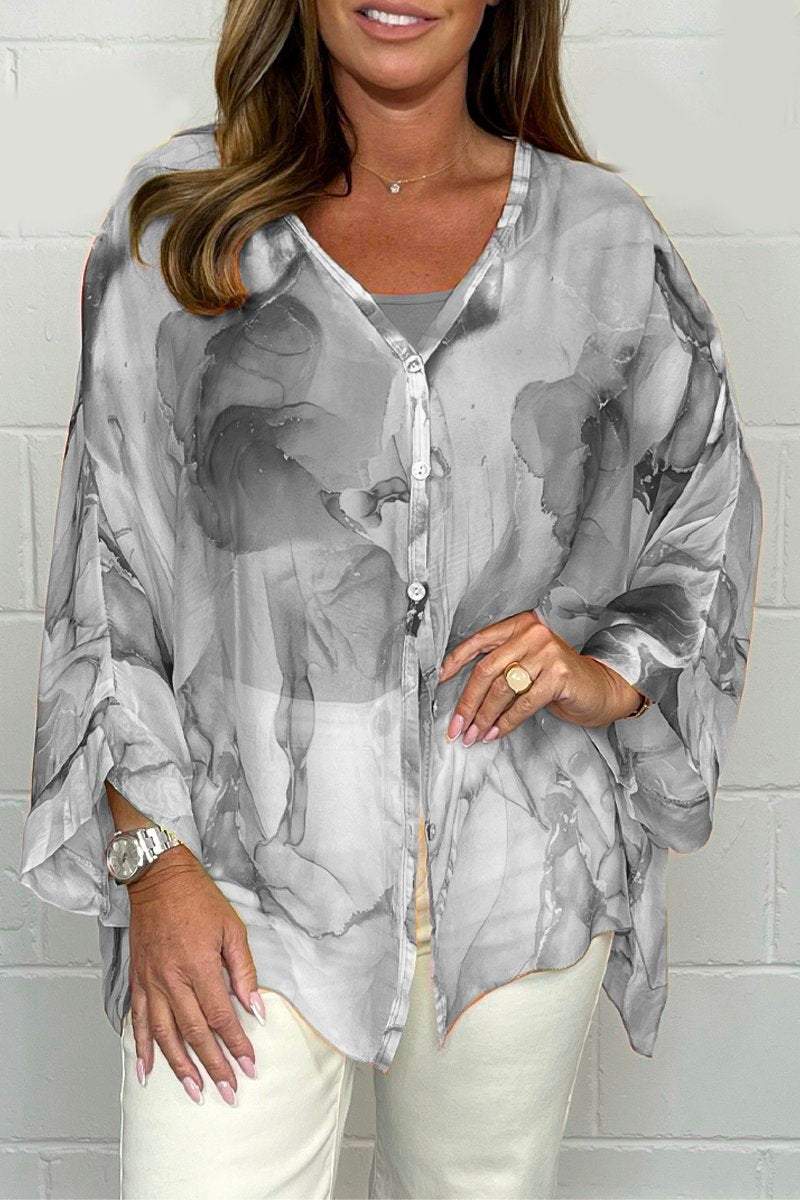 Femme portant une tunique fluide grise à motifs abstraits, manches longues, boutons, avec pantalon blanc. Mode élégante, style décontracté chic.