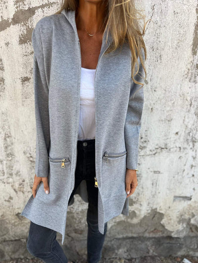 Femme portant un cardigan gris avec fermeture éclair, poches zippées, sur un mur texturé. Mode automne, style décontracté, tenue élégante.