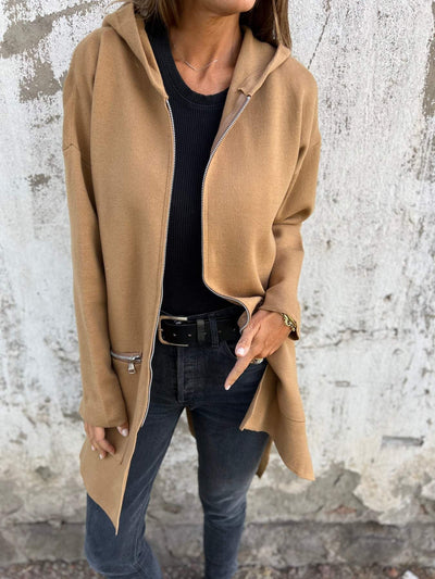 Femme portant un manteau camel à capuche, pull noir et jean gris, posant devant un mur texturé. Mode automne, style décontracté chic.