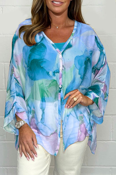 Femme portant une tunique fluide à motifs aquarelle bleu et vert, manches longues, sur fond blanc. Mode féminine élégante et colorée.