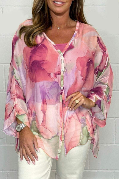 Blouse femme fluide à motifs floraux roses et violets, manches longues, portée avec un pantalon blanc. Mode élégante et tendance.