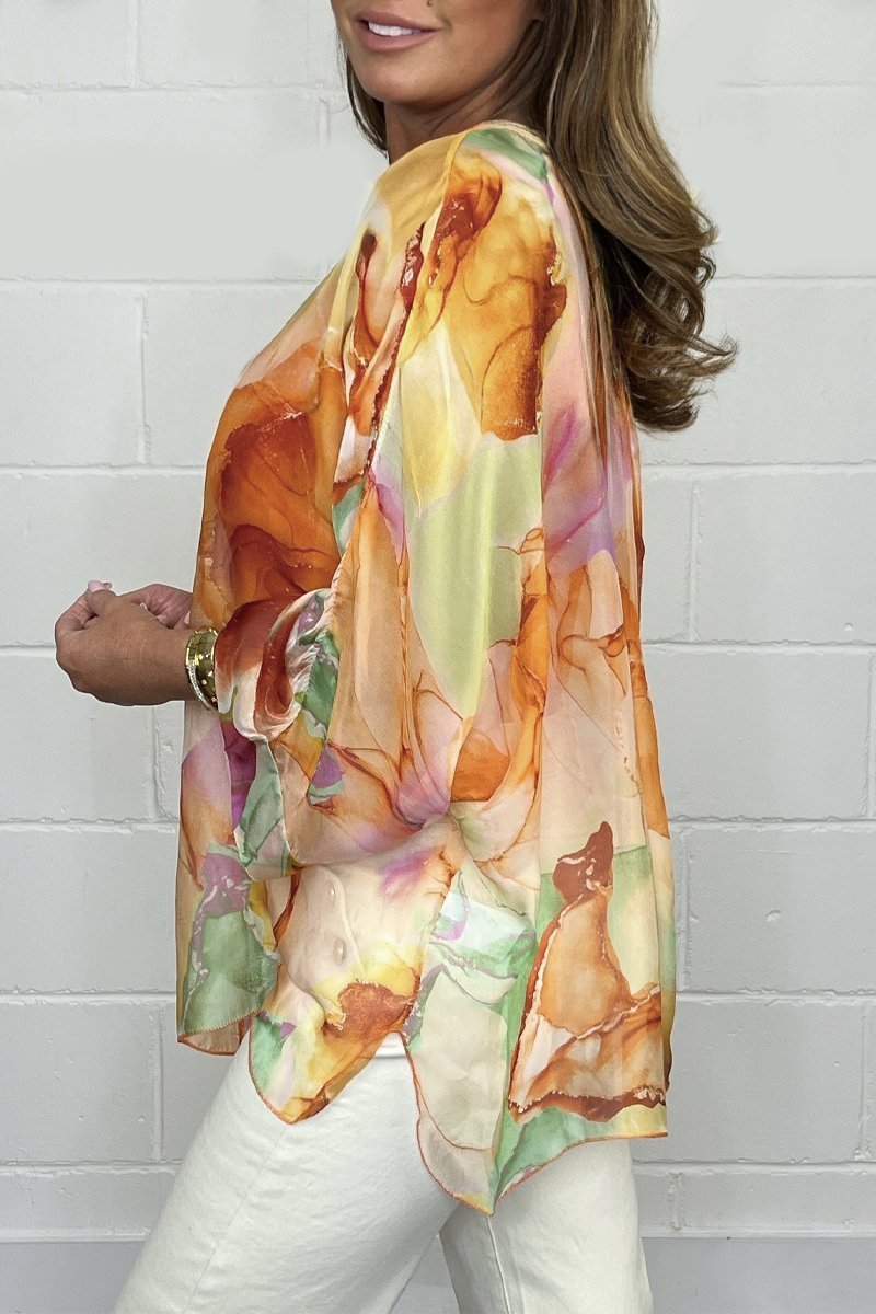 Blouse en soie colorée avec motifs floraux orange et rose, manches longues, portée par une femme aux cheveux bruns, mur blanc en arrière-plan.