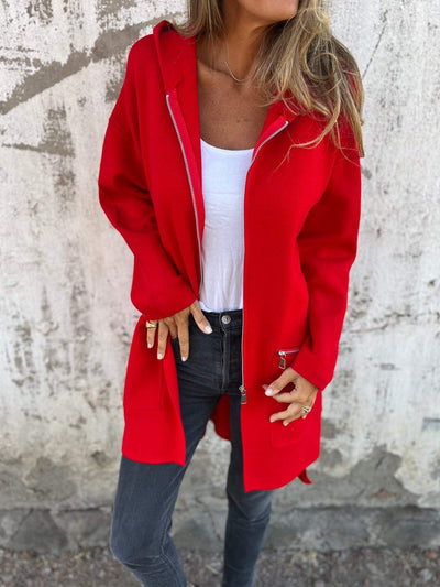 Femme portant un manteau rouge vif avec capuche, t-shirt blanc et jeans noirs, posant devant un mur texturé. Mode automne, style décontracté.