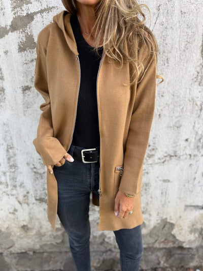 Femme portant un manteau camel à capuche, pull noir et jean gris, posant devant un mur texturé. Mode automne, style décontracté chic.