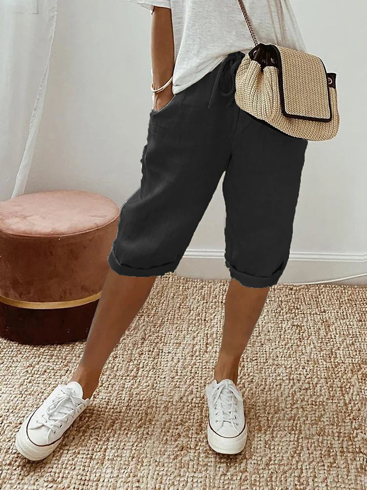 Femme portant un short noir décontracté, t-shirt blanc, baskets blanches et sac à main en paille, debout sur un tapis beige. Mode estivale chic.