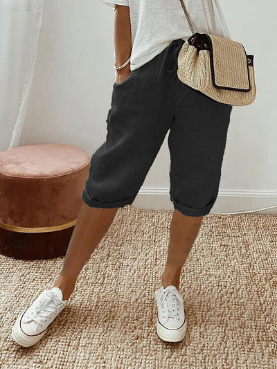 Femme portant un short noir décontracté, t-shirt blanc, baskets blanches et sac à main en paille, debout sur un tapis beige. Mode estivale chic.