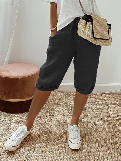 Femme en short noir décontracté, t-shirt blanc, sac à main en paille, baskets blanches, posant sur un tapis beige. Mode estivale chic et confortable.