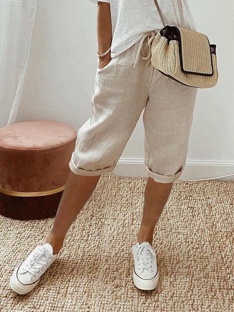 Personne portant un short en lin beige, t-shirt blanc, baskets blanches et sac à main en paille, debout sur un tapis en jute. Mode décontractée estivale.