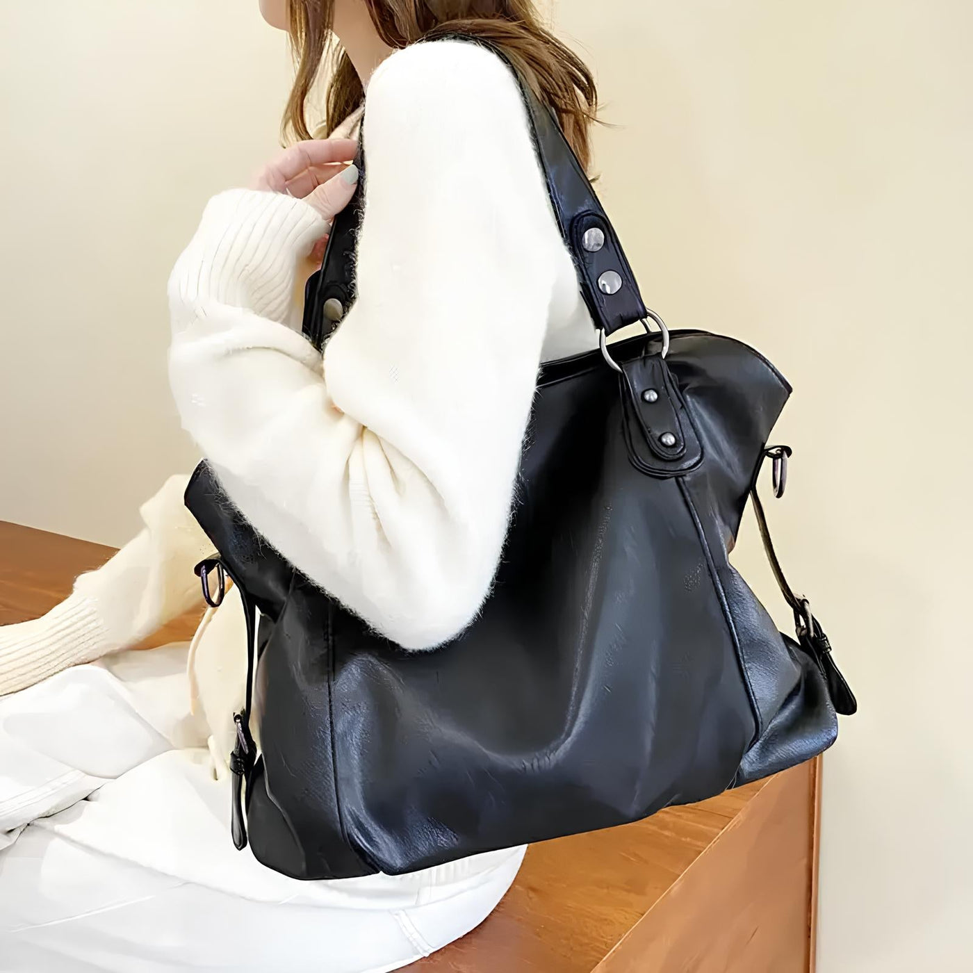 Sac à main en cuir noir porté par une femme en pull blanc, posé sur une table en bois. Mode féminine, accessoire élégant, tendance automne.