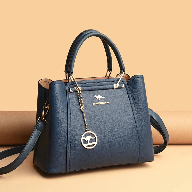 Sac à main bleu élégant en cuir avec bandoulière, logo doré et pendentif, posé sur fond beige. Accessoire mode femme chic et tendance.