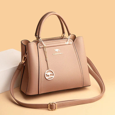 Sac à main en cuir beige élégant avec bandoulière, détails dorés et pendentif logo, posé sur fond beige. Accessoire mode femme chic et tendance.