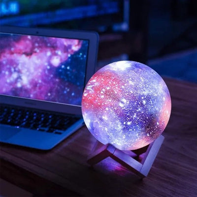 Lampe galaxie LED colorée sur support en bois, posée à côté d'un ordinateur portable affichant une image de l'espace, éclairage d'ambiance cosmique.