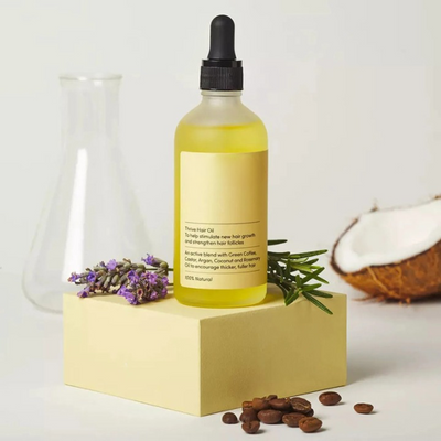 Flacon d'huile capillaire sur socle, entouré de lavande, romarin, noix de coco et grains de café. Produit naturel pour croissance et soin des cheveux.