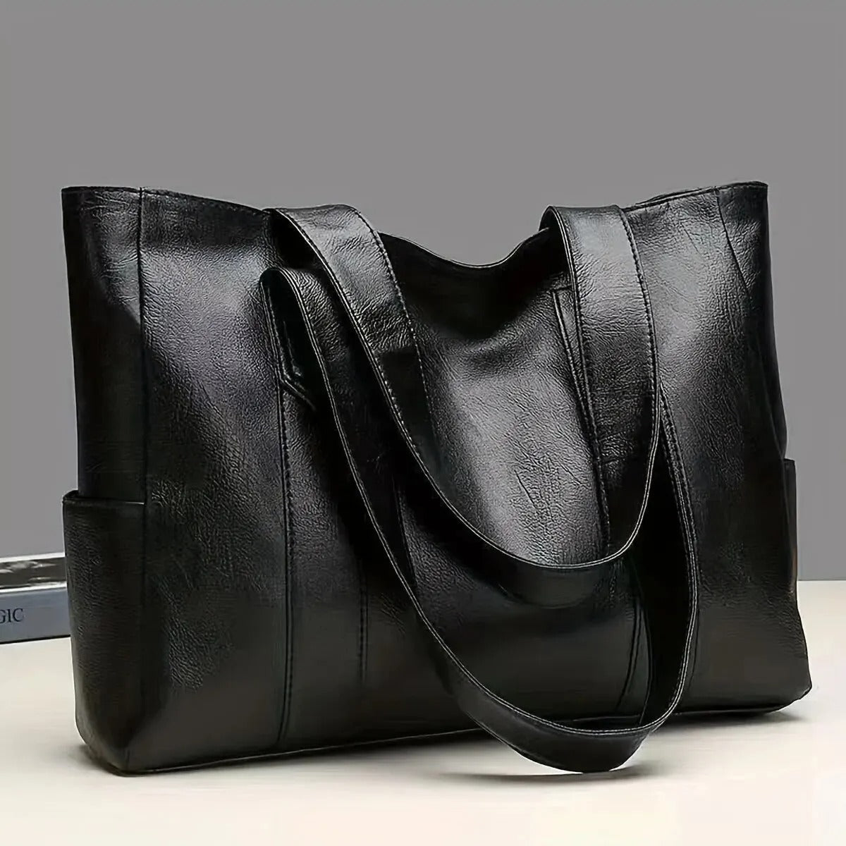 Sac à main en cuir noir élégant avec anses longues, posé sur une surface blanche. Accessoire mode femme, design moderne et spacieux.