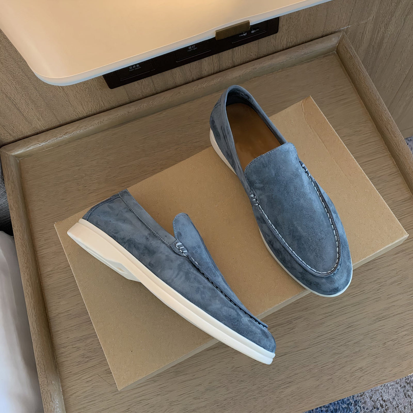 Mocassins en daim bleu sur boîte en carton, design élégant et moderne, semelle blanche contrastante, posés sur table en bois.