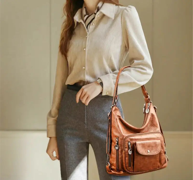 Femme portant un sac à main en cuir marron avec poches zippées, tenue élégante avec chemisier beige et pantalon gris, mode chic et moderne.