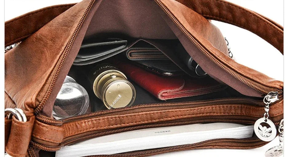 Sac à main en cuir marron ouvert, contenant portefeuille, rouge à lèvres, flacons de parfum, et cahier. Accessoire mode femme, rangement pratique.