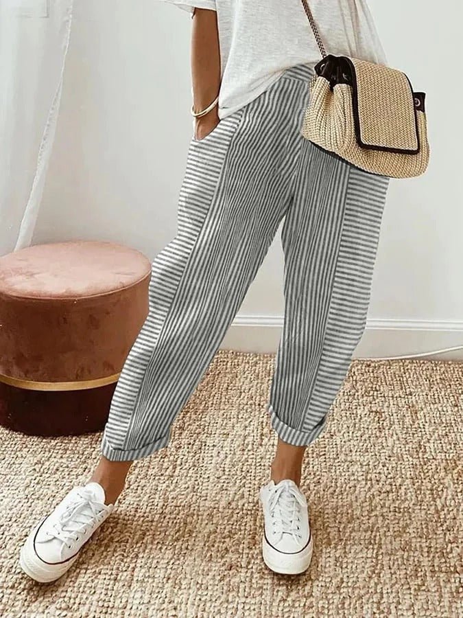 Femme portant un pantalon rayé gris et blanc, t-shirt blanc, baskets blanches, sac en osier, posant sur un tapis beige. Mode décontractée chic.