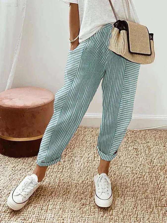 Personne portant un pantalon rayé bleu et blanc, t-shirt blanc, baskets blanches, sac en osier, debout sur un tapis beige. Mode décontractée chic.