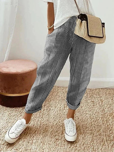 Femme portant un pantalon rayé bleu et blanc, t-shirt blanc, baskets blanches, sac en osier, debout sur un tapis beige. Mode décontractée chic.