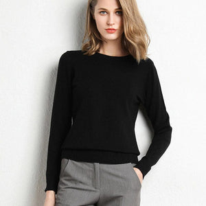 Femme portant un pull noir élégant et un pantalon gris, posant contre un mur blanc. Mode féminine chic, tenue automne-hiver tendance.
