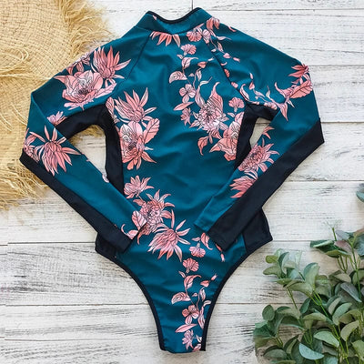 Maillot de bain une pièce à manches longues, motif floral rose sur fond bleu, posé sur un sol en bois avec des plantes vertes en arrière-plan.