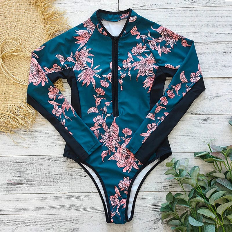 Maillot de bain une pièce à manches longues, motif floral rose sur fond bleu, posé sur un fond en bois clair avec des éléments naturels.