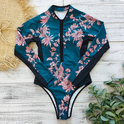 Maillot de bain une pièce à manches longues, motif floral rose sur fond bleu, posé sur un fond en bois clair avec des éléments naturels.