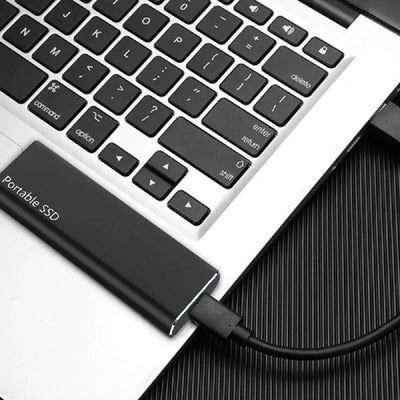 Disque SSD portable connecté à un ordinateur portable via un câble USB, posé sur un clavier argenté. Stockage externe rapide et compact.