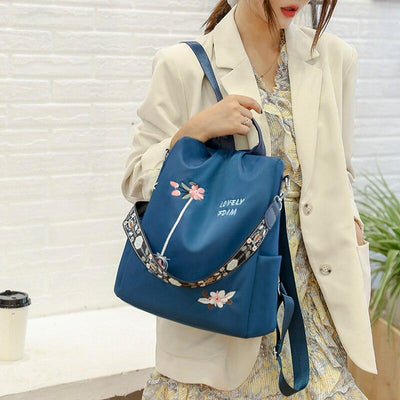 Femme portant un sac à dos bleu avec motifs floraux, tenue élégante beige, mur en briques blanches en arrière-plan. Mode féminine chic.