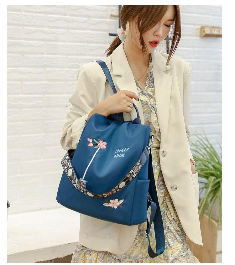 Femme portant un sac à dos bleu avec motifs floraux, tenue élégante beige, mur blanc en arrière-plan. Mode féminine, accessoires tendance.