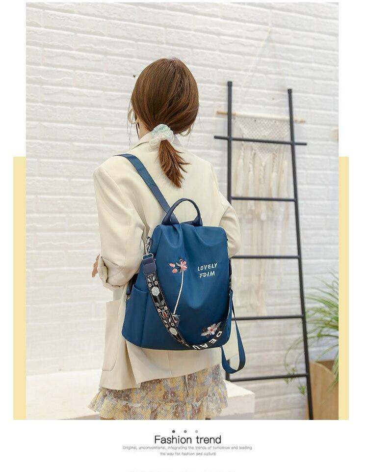 Femme de dos portant un sac à dos bleu avec motif floral, veste beige, robe à volants, devant un mur blanc et une échelle décorative.