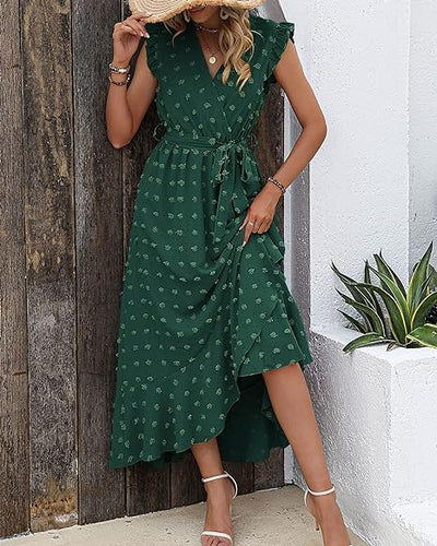 Robe longue verte à motifs floraux, sans manches, avec ceinture, portée par une femme tenant un chapeau de paille, posant près d'un mur blanc.