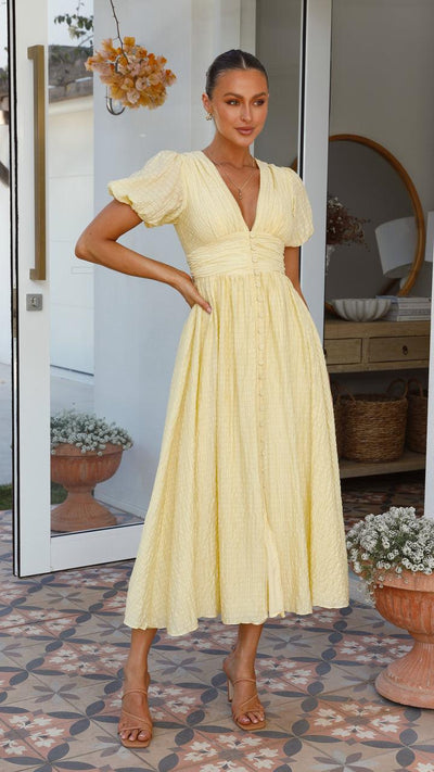 Femme en robe jaune élégante, debout sur un patio carrelé, porte des sandales marron. Décor floral et miroir en arrière-plan. Mode estivale chic.