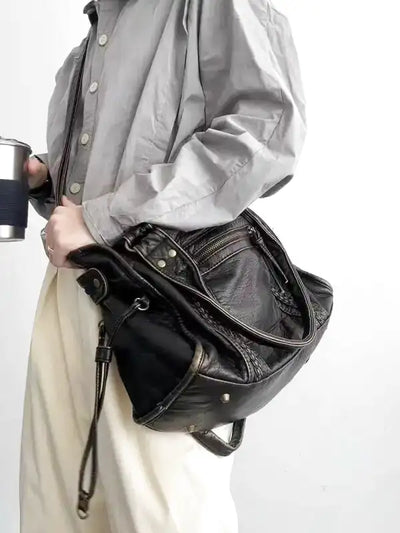 Personne portant un sac à main en cuir noir avec des détails métalliques, tenue décontractée avec chemise grise et pantalon blanc, tenant une tasse.