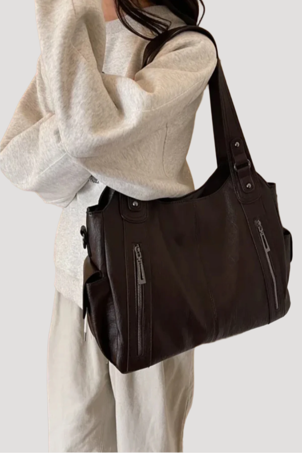 Femme portant un sac à main en cuir noir avec fermetures éclair, tenue décontractée en sweat gris et pantalon beige. Mode, accessoires, élégance.