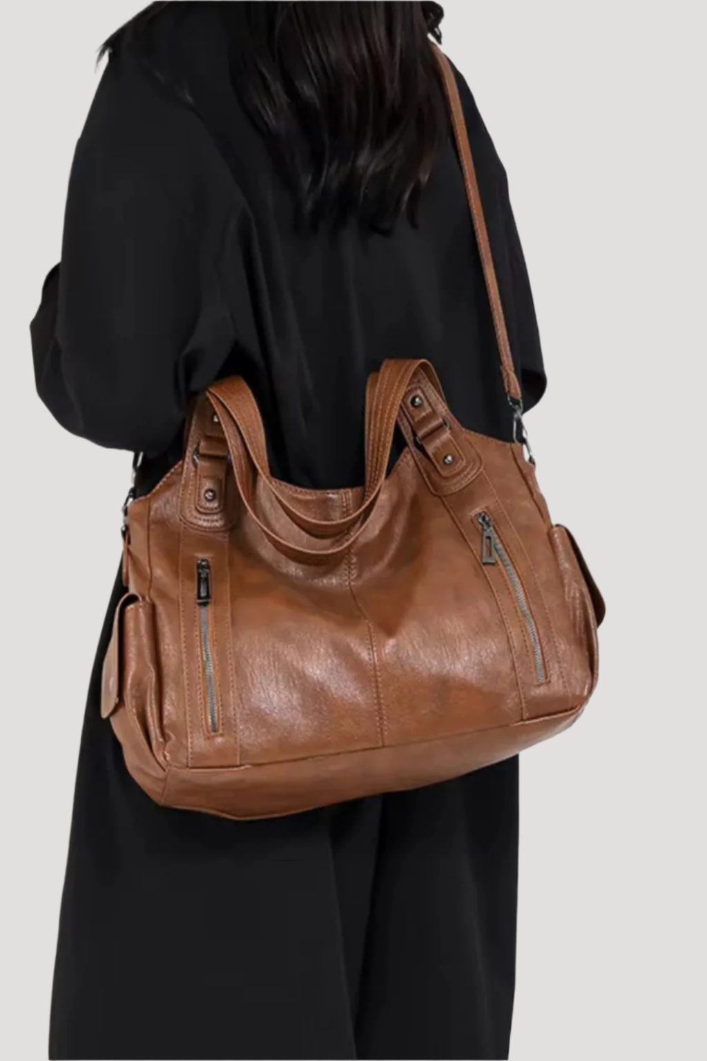 Sac à main en cuir marron porté par une personne en manteau noir, avec fermetures éclair et anses robustes. Accessoire mode élégant et pratique.