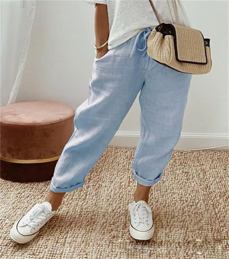 Femme portant un pantalon en lin bleu clair, t-shirt blanc, baskets blanches et sac en osier, posant sur un tapis beige. Mode décontractée chic.