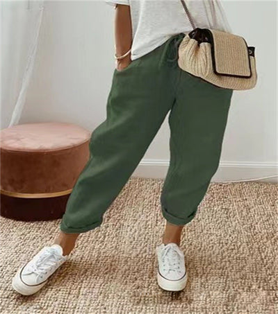 Femme portant un pantalon vert décontracté, t-shirt blanc, baskets blanches et sac à main beige, posant sur un tapis beige. Mode décontractée chic.