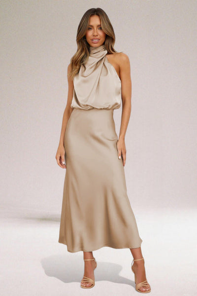 Femme portant une robe longue beige élégante, sans manches, avec col drapé, posant sur fond neutre. Mode féminine chic, tenue de soirée.