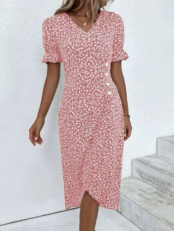 Robe midi fleurie rose à manches bouffantes, col en V, boutonnée, portée par une femme sur fond de mur blanc et escaliers. Mode féminine élégante.